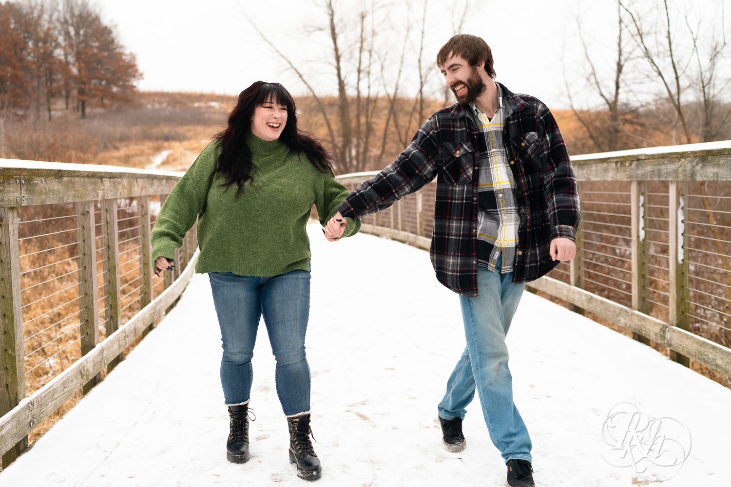 Man in flannel and woman in green sweater and jeans laugh and walk across snowy bridge at Lebanon Hills in Eagan, Minnesota.