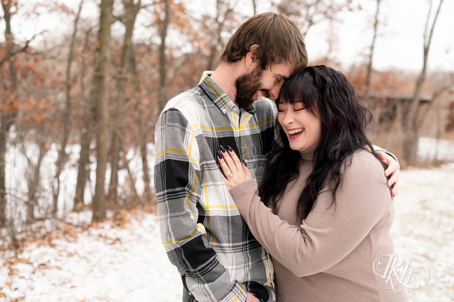 Man in flannel and woman in brown dress smile during winter engagement photos at Lebanon Hills Regional Park in Eagan, Minnesota.