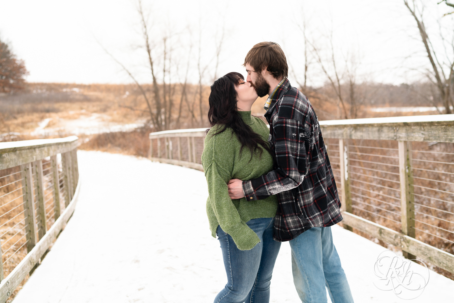 Man in flannel and woman in green sweater and jeans kiss on snowy bridge at Lebanon Hills in Eagan, Minnesota.