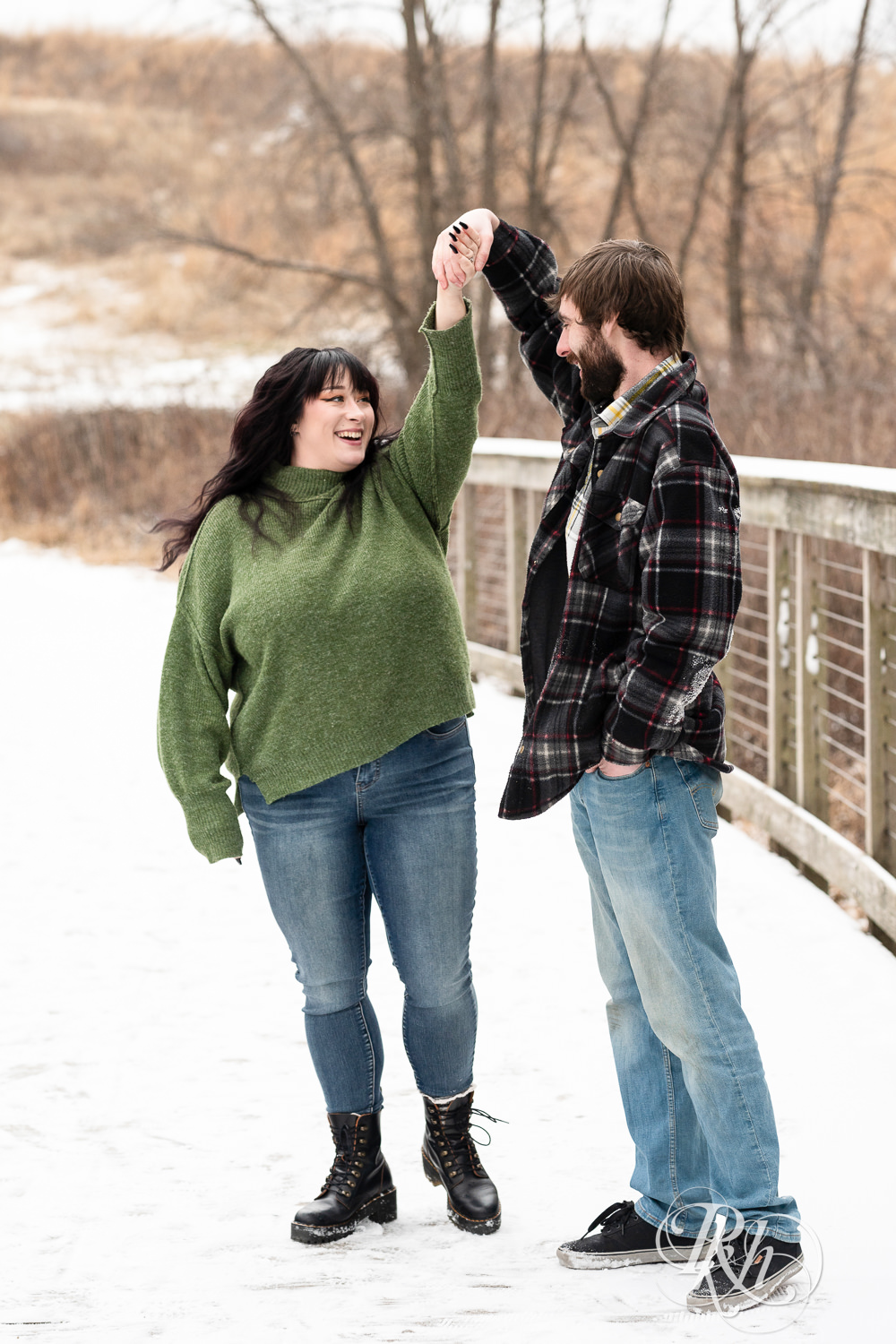 Man in flannel and woman in green sweater and jeans dance snowy bridge at Lebanon Hills in Eagan, Minnesota.
