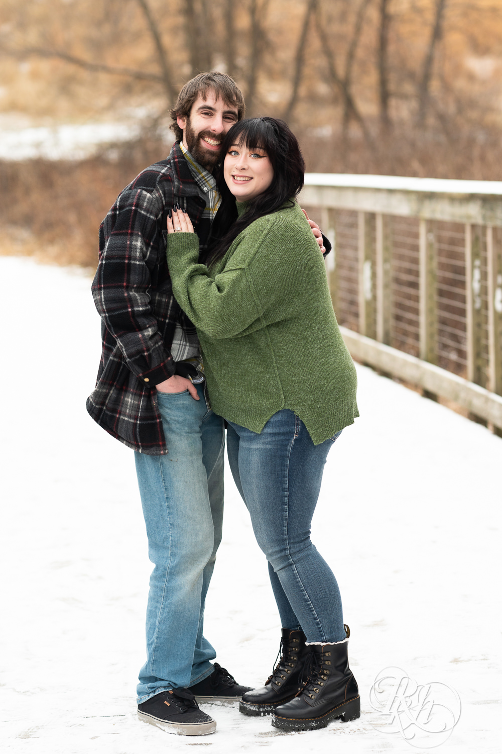Man in flannel and woman in green sweater and jeans smile on snowy bridge at Lebanon Hills in Eagan, Minnesota.