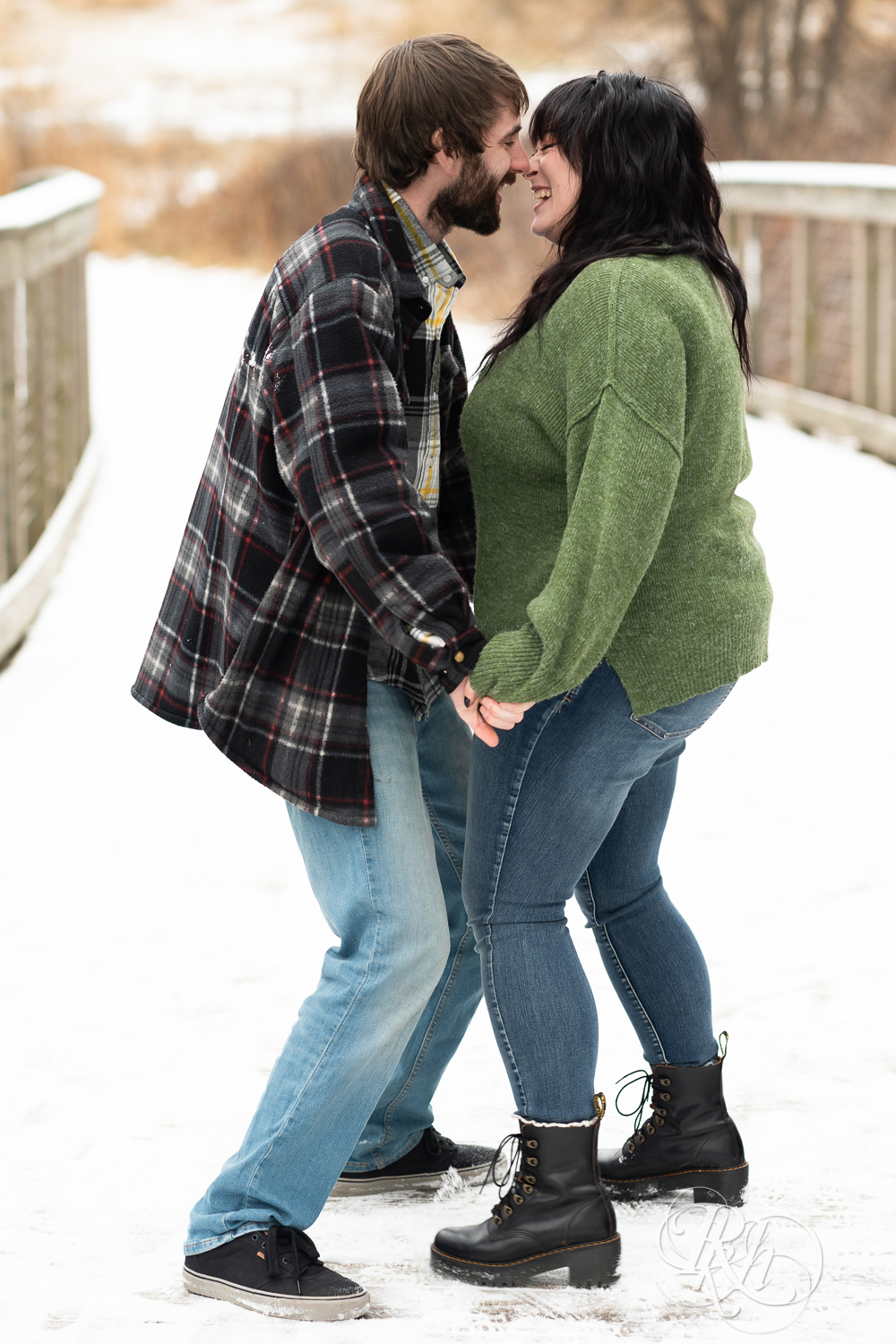 Man in flannel and woman in green sweater and jeans laugh on snowy bridge at Lebanon Hills in Eagan, Minnesota.