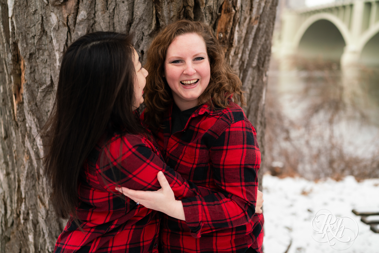 Red headed woman and Asian woman in red flannels laugh during winter engagement photography in Minneapolis, Minnesota.
