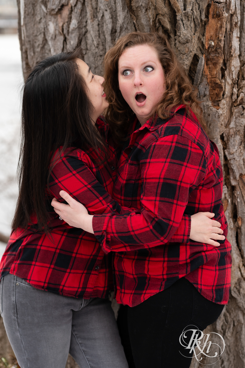 Red headed woman and Asian woman in red flannels laugh during winter engagement photography in Minneapolis, Minnesota.