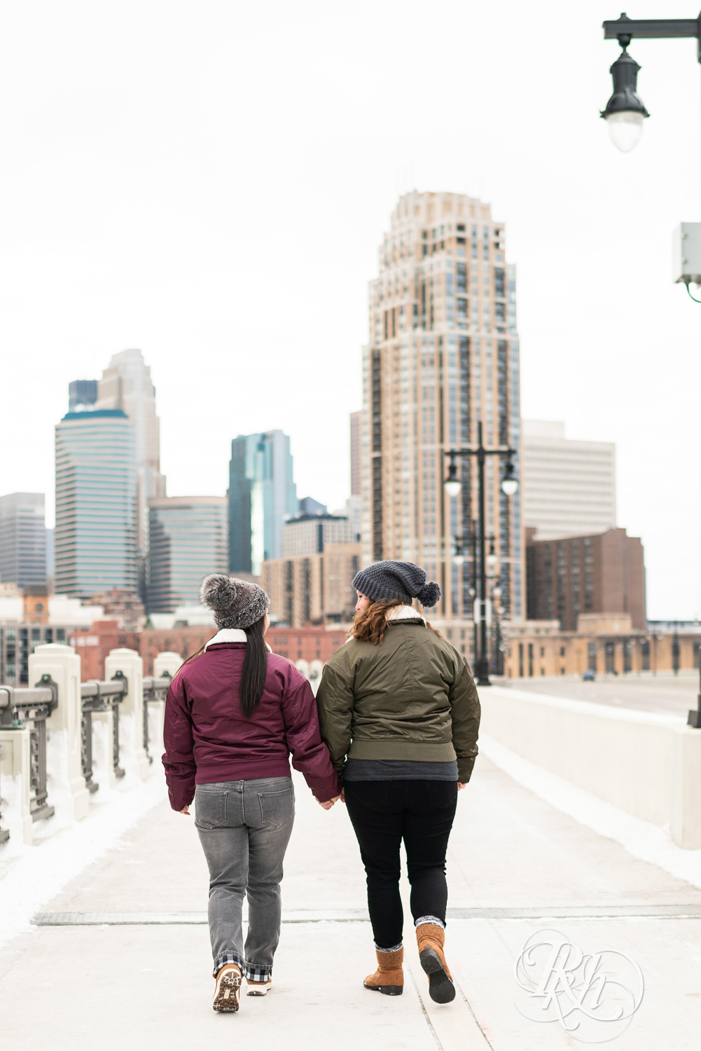 Red headed woman and Asian woman in jackets walk down Third Street Bridge during winter engagement photography in Minneapolis, Minnesota.
