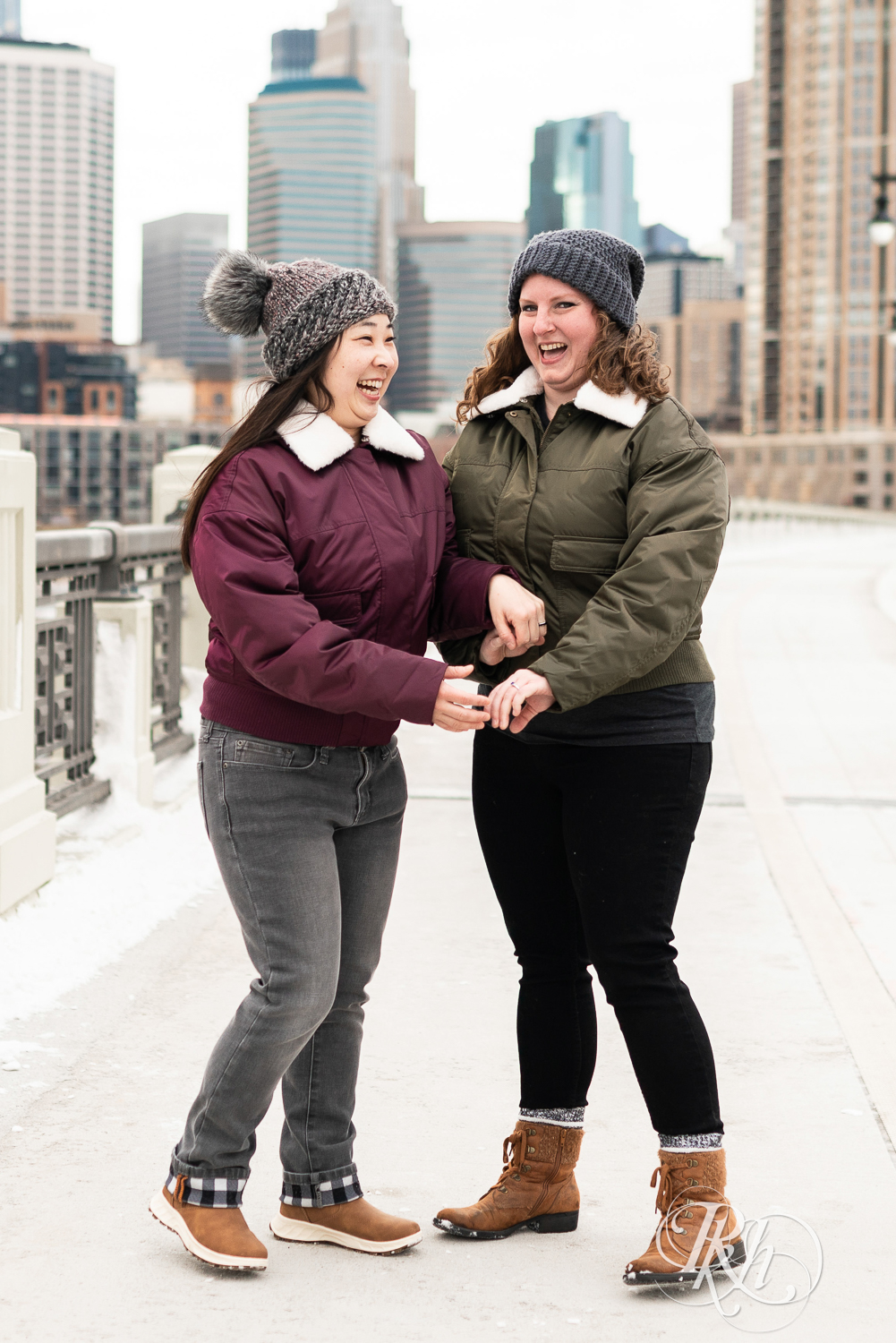 Red headed woman and Asian woman in jackets walk down Third Street Bridge during winter engagement photography in Minneapolis, Minnesota.
