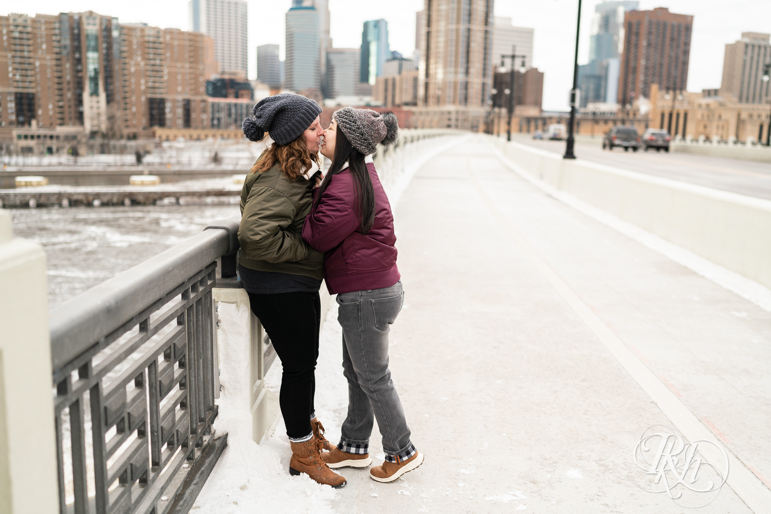 Red headed woman and Asian woman in jackets kiss on Third Street Bridge during winter engagement photography in Minneapolis, Minnesota.