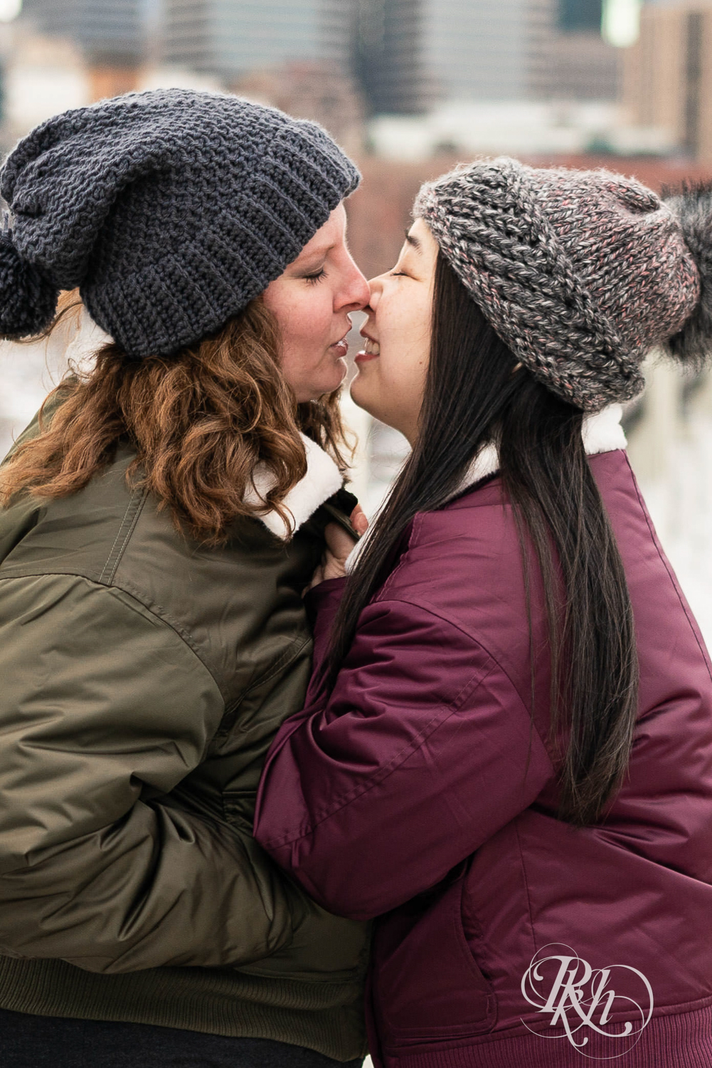 Red headed woman and Asian woman in jackets kiss on Third Street Bridge during winter engagement photography in Minneapolis, Minnesota.