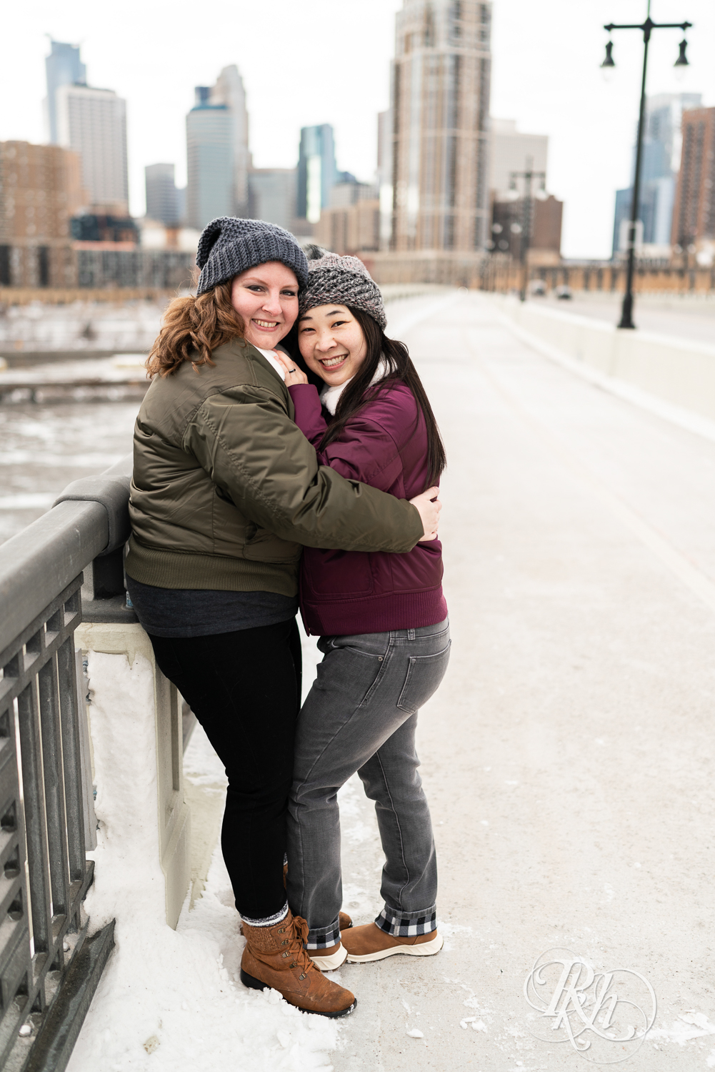 Red headed woman and Asian woman in jackets snuggle on Third Street Bridge during winter engagement photography in Minneapolis, Minnesota.