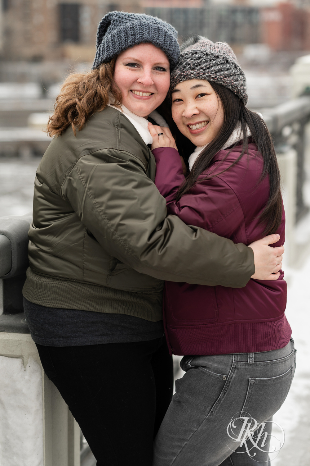 Red headed woman and Asian woman in jackets snuggle on bridge during winter engagement photography in Minneapolis, Minnesota.