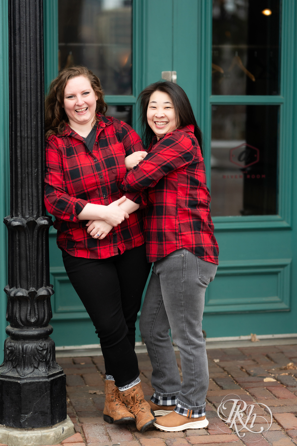 Red headed woman and Asian woman in red flannels smile during winter engagement photography in Minneapolis, Minnesota.