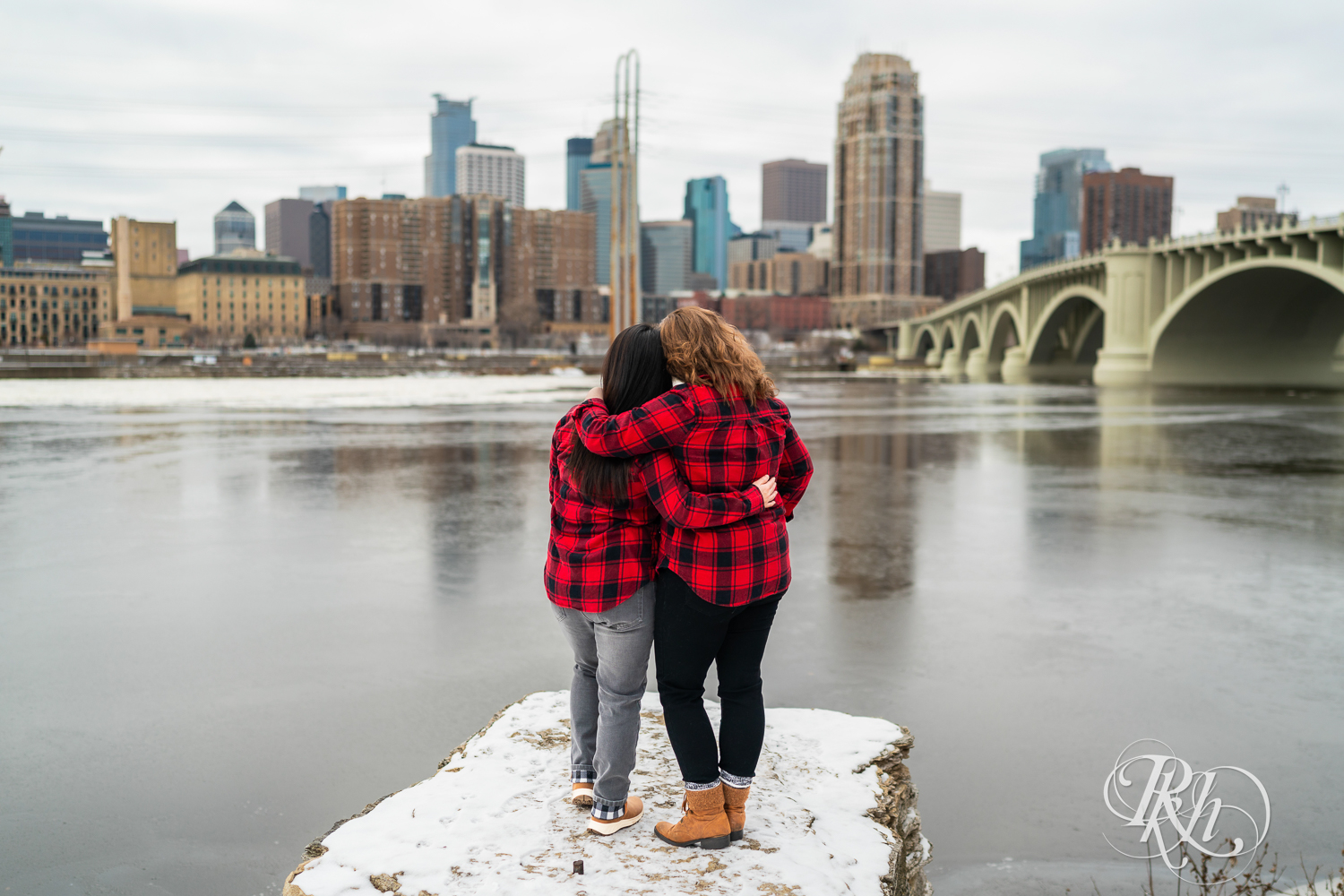 Red headed woman and Asian woman in red flannels smile during winter engagement photography in Minneapolis, Minnesota in front of the Mississippi River.
