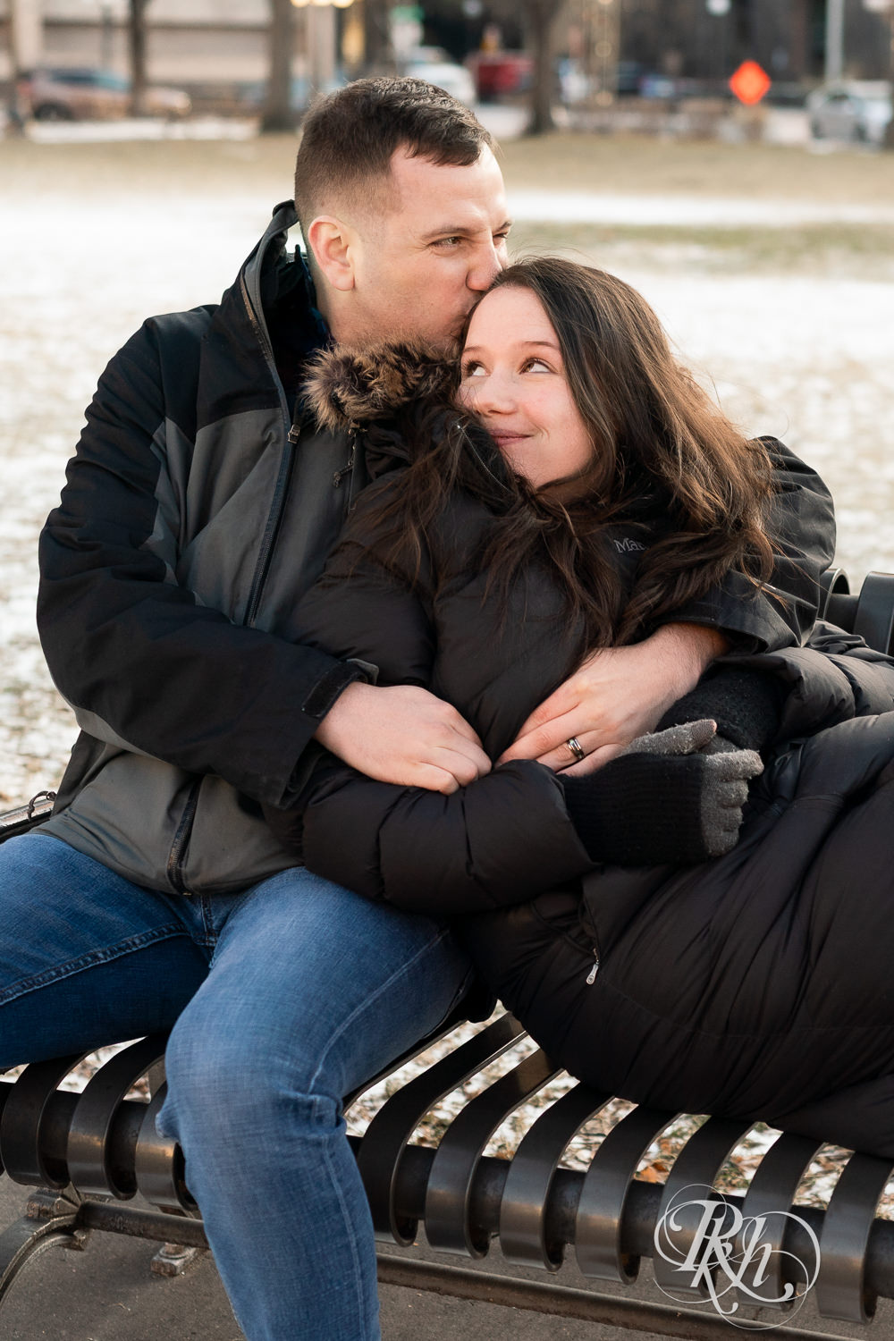 Man and woman in jackets snuggle on park bench in Saint Paul, Minnesota during winter engagement session.