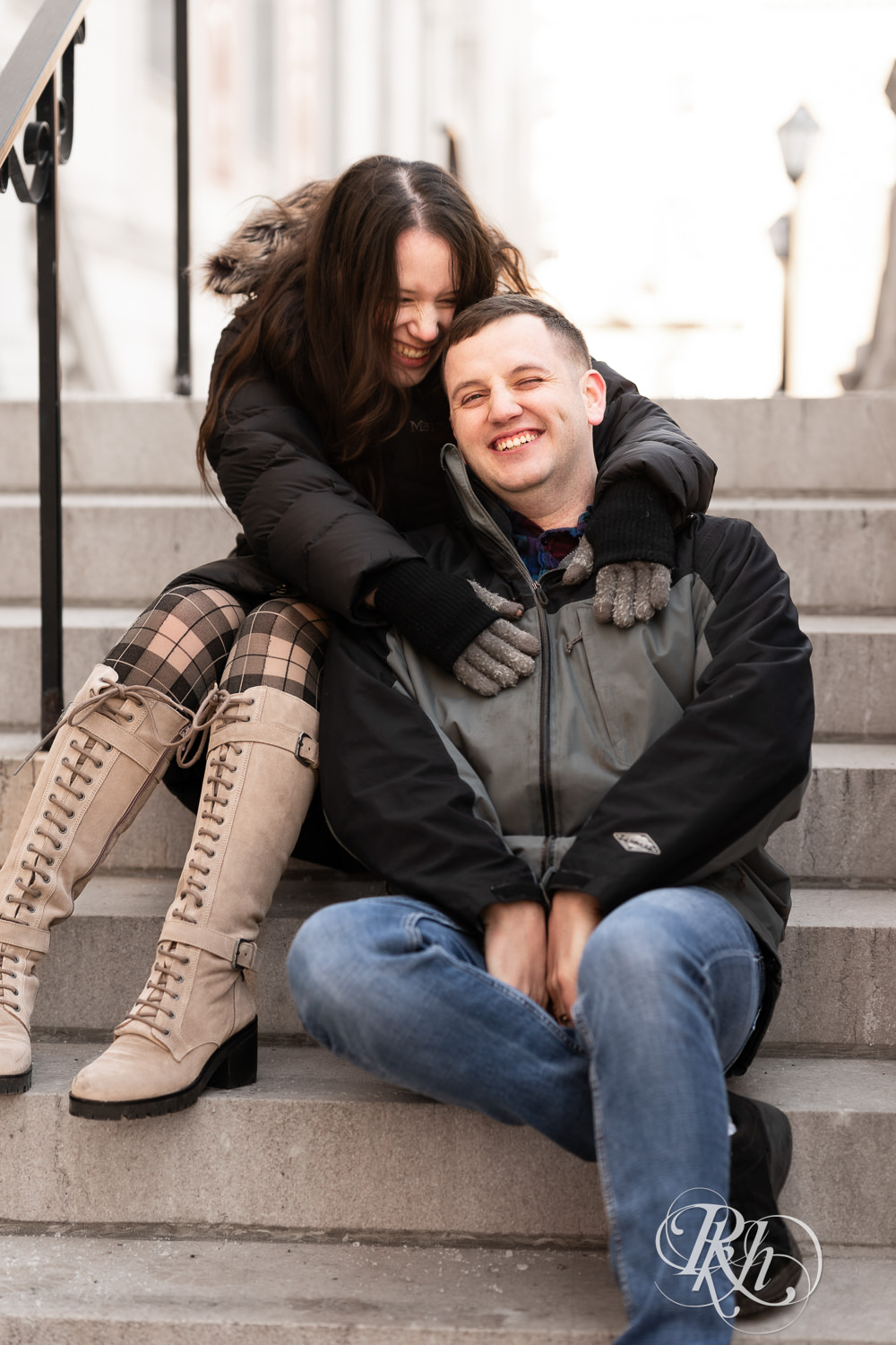 Man and woman in jackets snuggle on stairs in Saint Paul, Minnesota during winter engagement session.