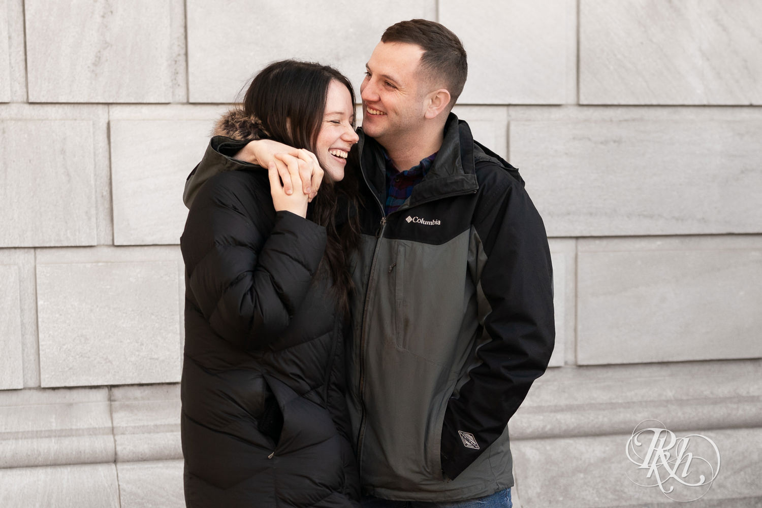Man and woman in jackets snuggle and kiss in front of the Saint Paul Library in Saint Paul, Minnesota during winter engagement session.