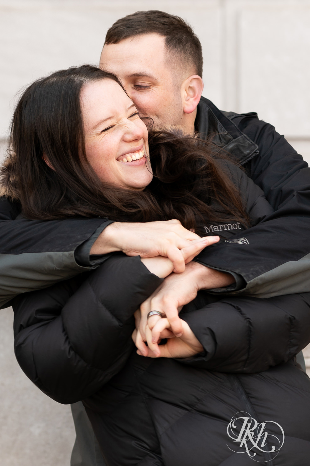 Man and woman in jackets snuggle and kiss in front of the Saint Paul Library in Saint Paul, Minnesota during winter engagement session.