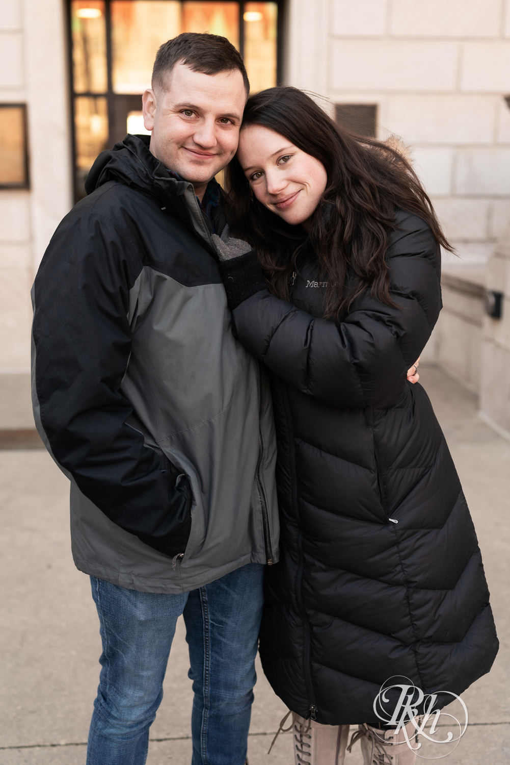 Man and woman in jackets snuggle and smile in front of the Saint Paul Library in Saint Paul, Minnesota during winter engagement session.