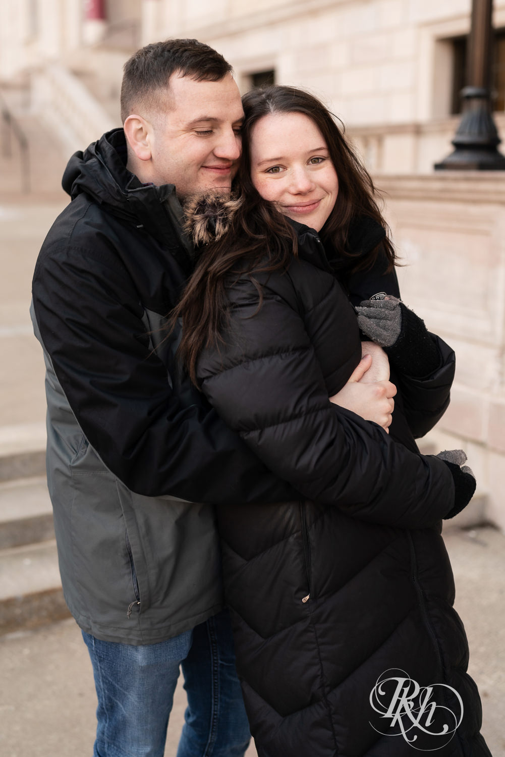 Man and woman in jackets snuggle in front of the Saint Paul Library in Saint Paul, Minnesota during winter engagement session.