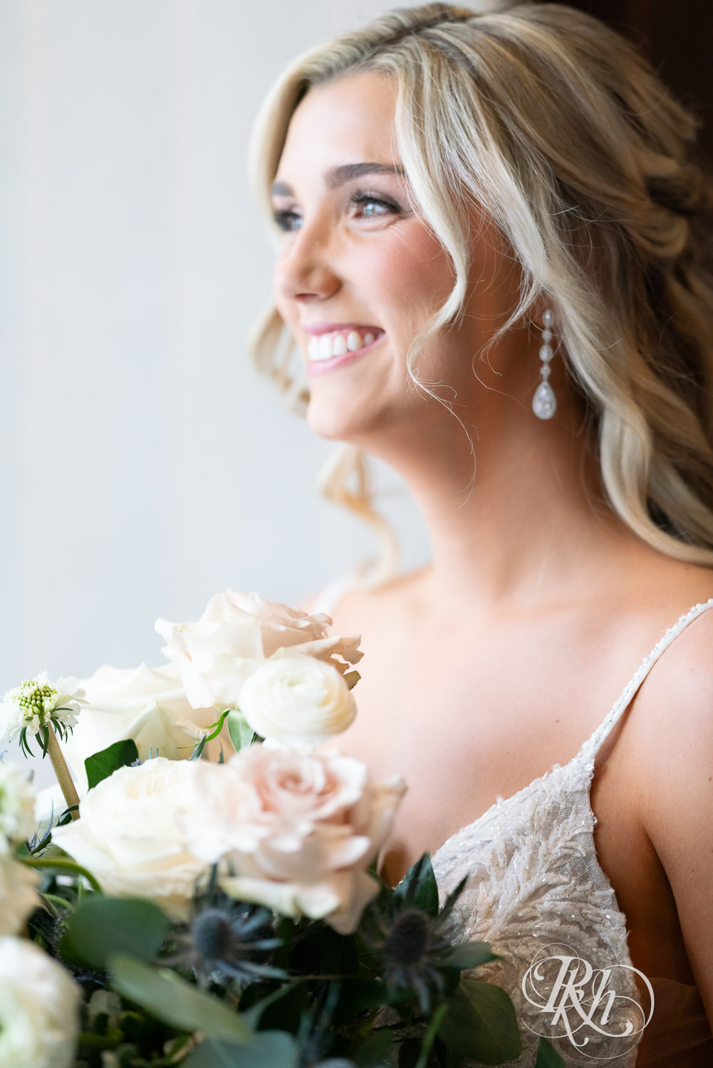 Bride smiles holding flowers in front of window on wedding day at Bavaria Downs in Chaska, Minnesota.