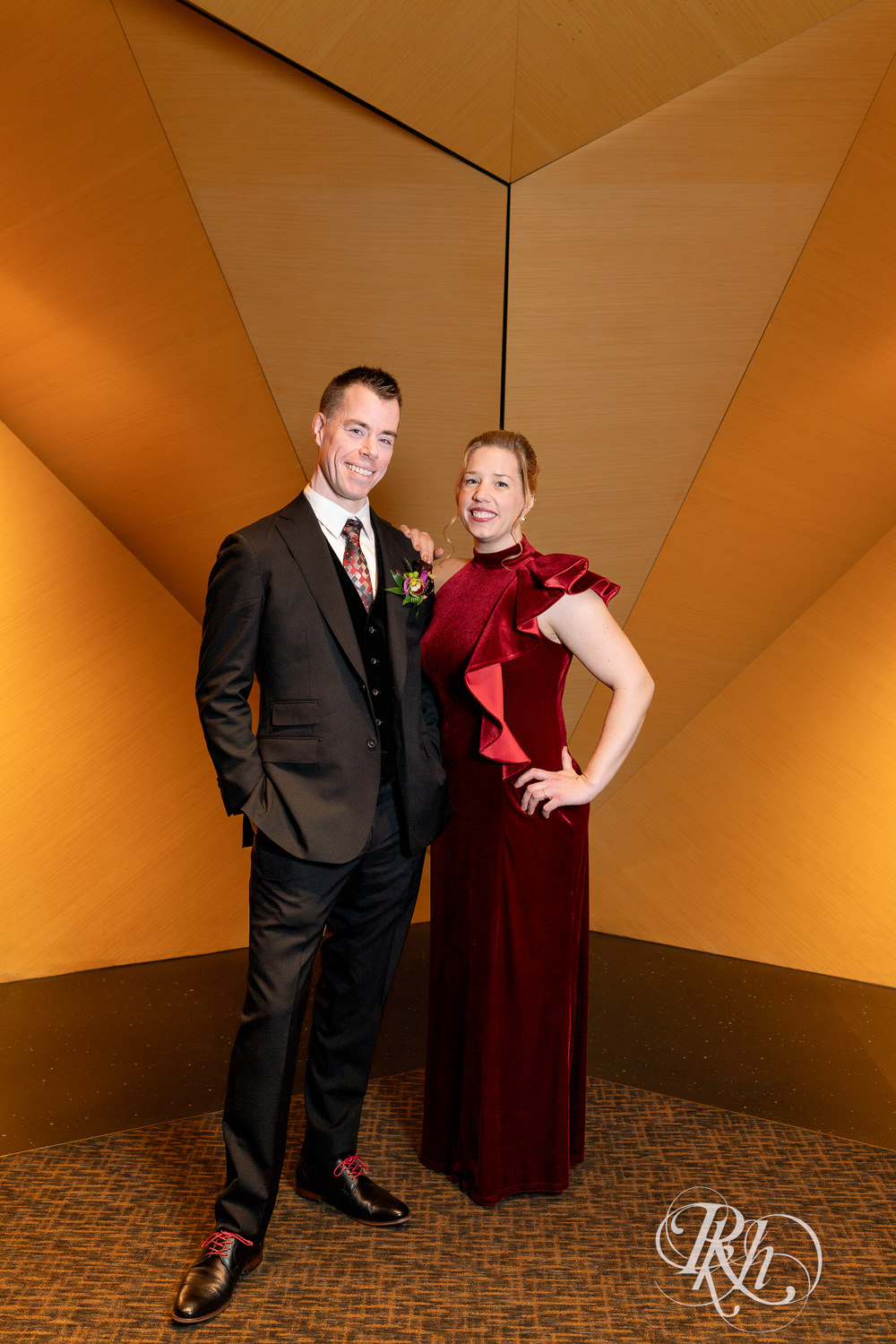 Bride wearing a red colorful wedding dress and groom in a black suit stand in front of a gold wall.