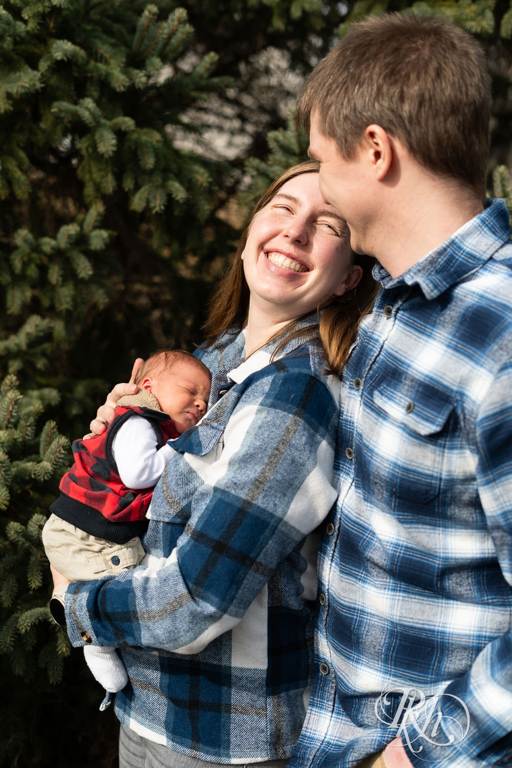Man and woman dressed in flannel hold their newborn in front of pine trees in Ham Lake, Minnesota.