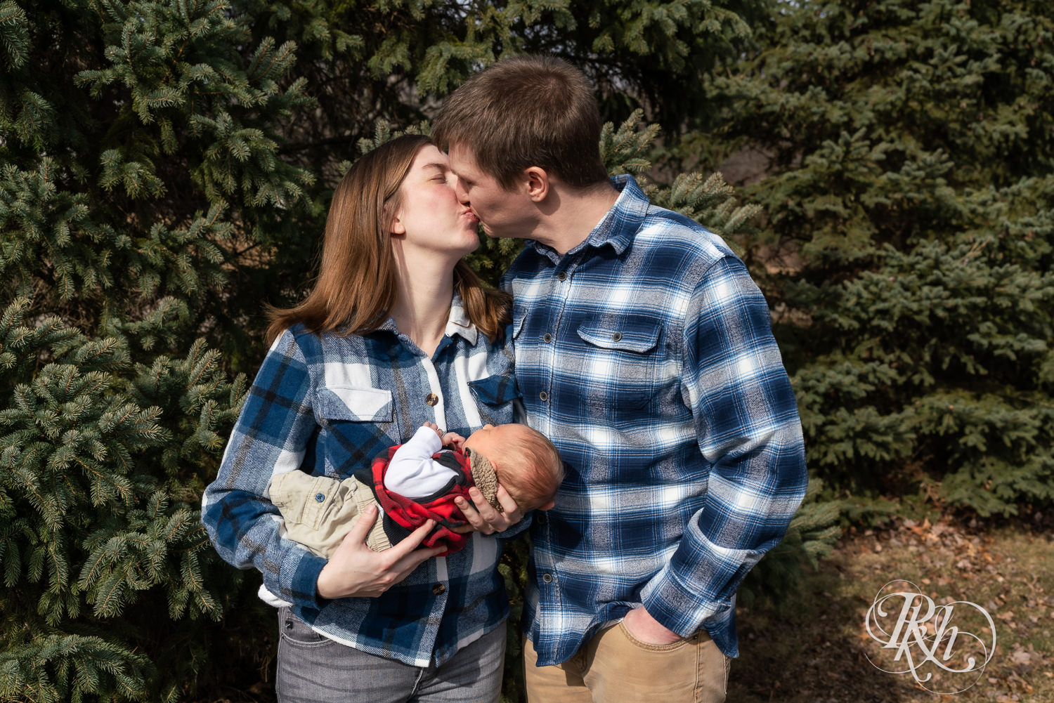 Man and woman dressed in flannel hold their newborn in front of pine trees in Ham Lake, Minnesota.