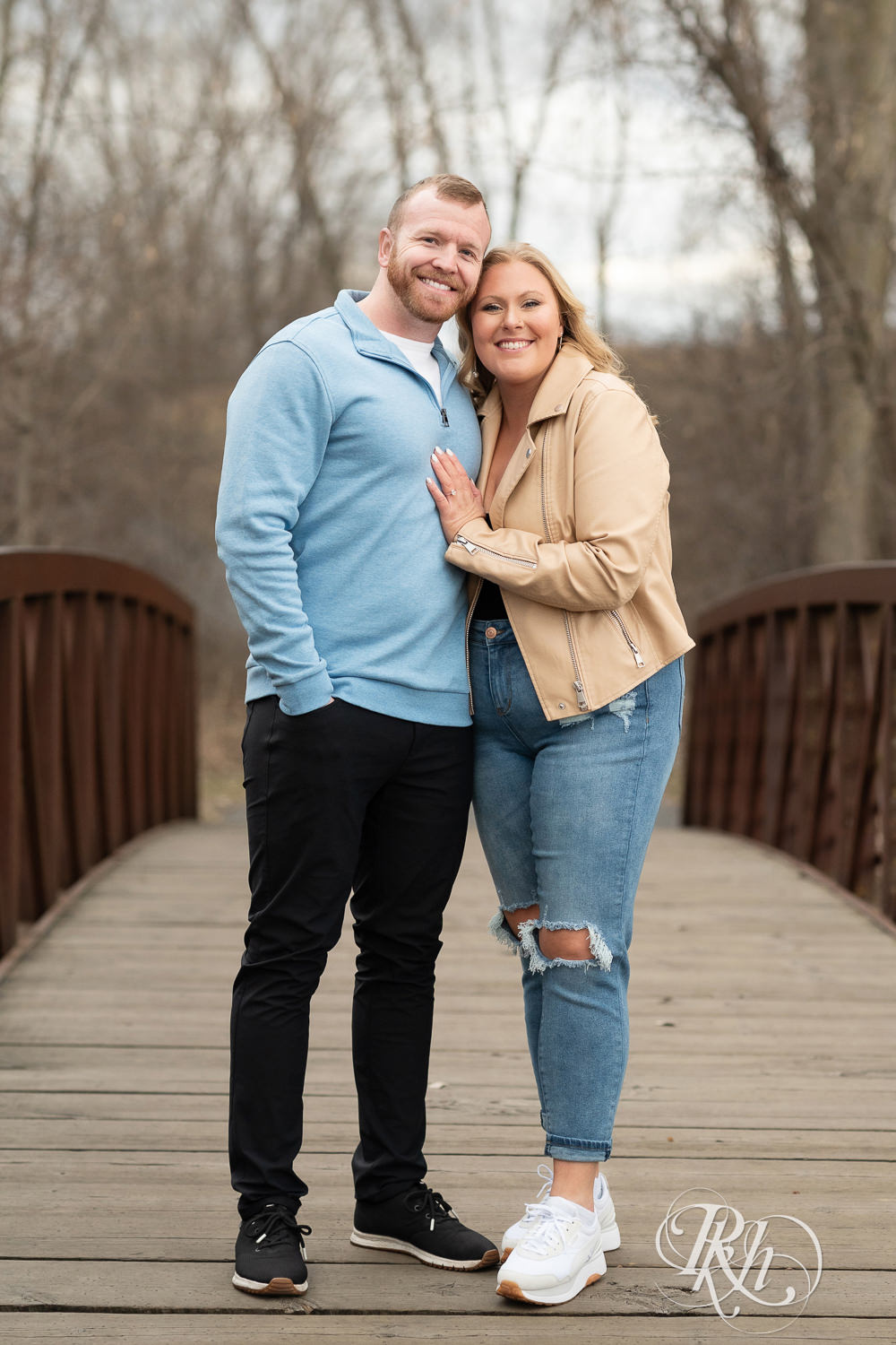 Man in blue shirt and woman in jeans smile on bridge during engagement photography at Rice Creek North Regional Trail in Shoreview, Minnesota.