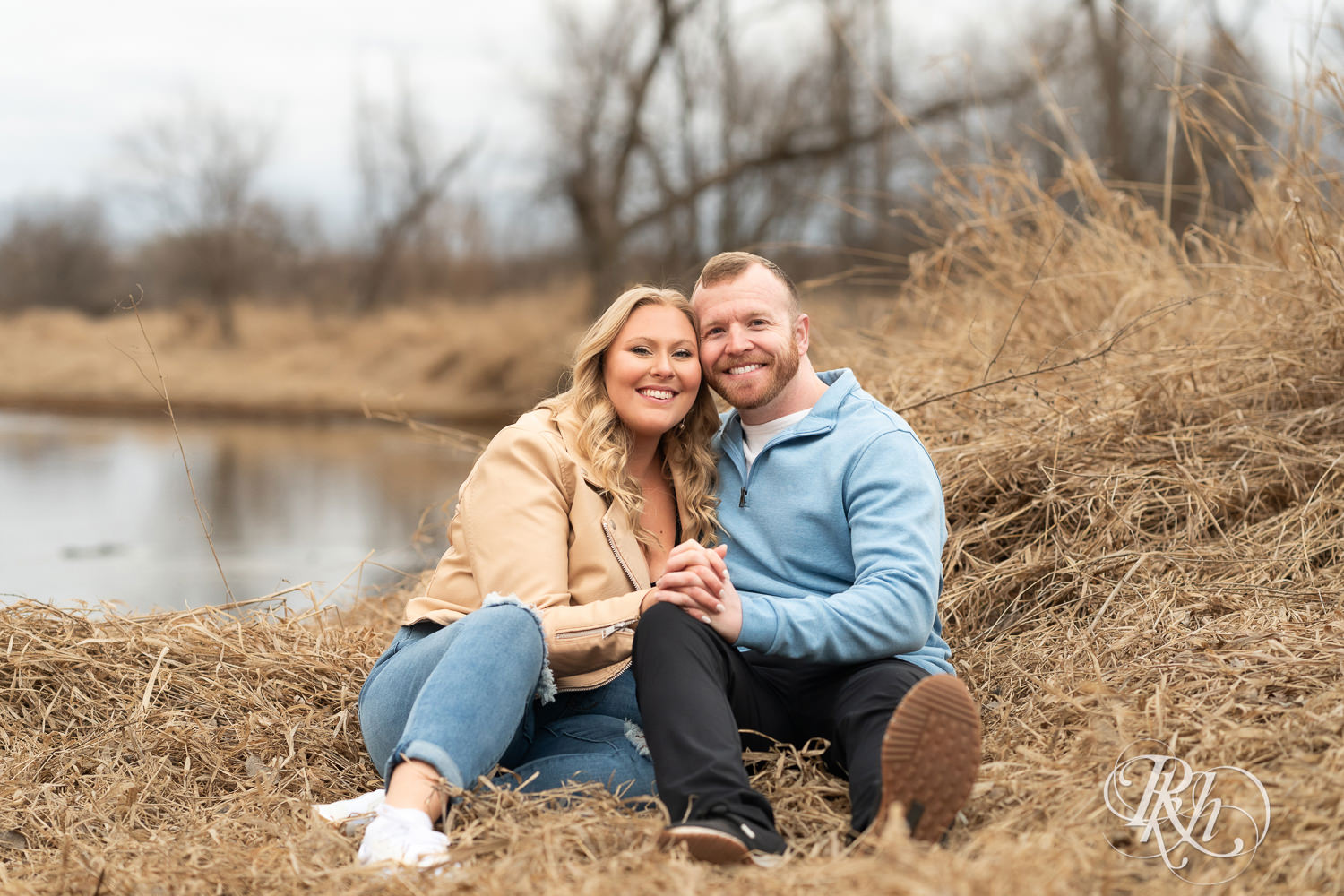 Man in blue shirt and woman in jeans smile on creekbed during engagement photography at Rice Creek Regional Trail in Shoreview, Minnesota.