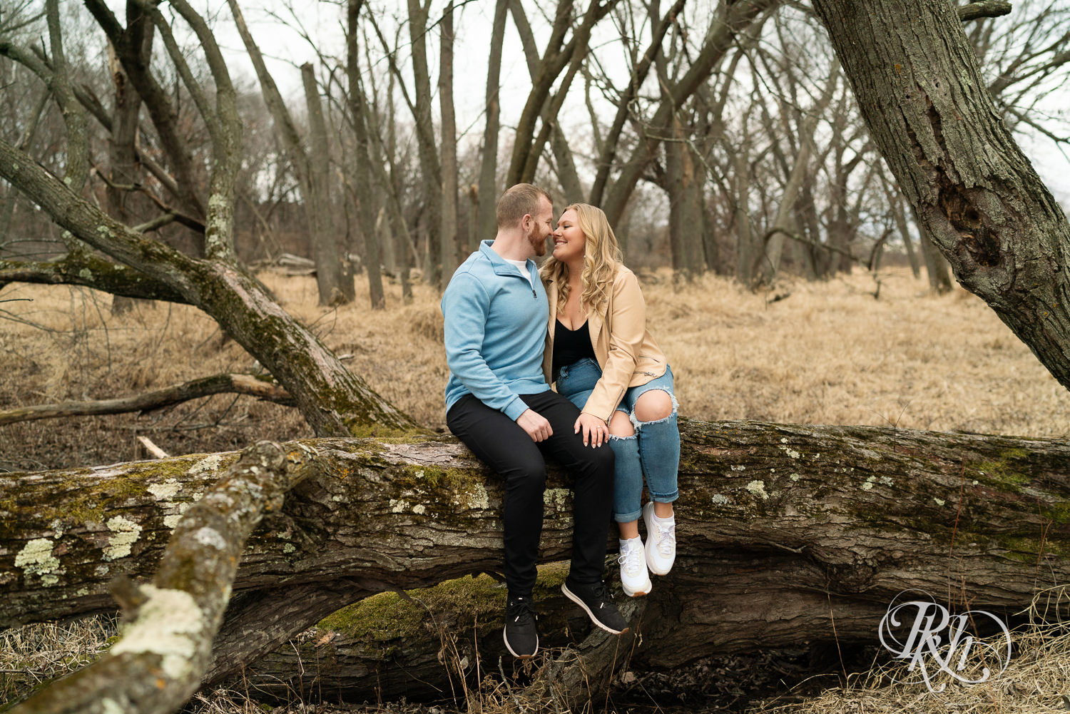 Man in blue shirt and woman in jeans sit on fallen tree during engagement photography at Rice Creek Regional Trail in Shoreview, Minnesota.