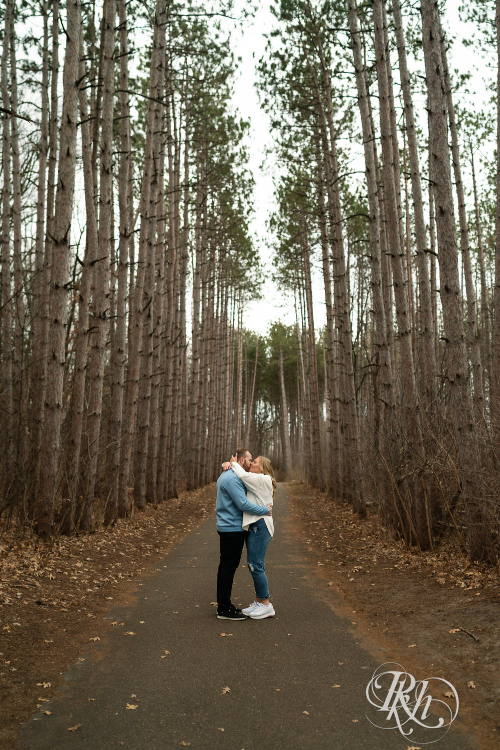 Man in blue shirt and woman in jeans kiss between tall trees during engagement photography at Rice Creek Regional Trail in Shoreview, Minnesota.
