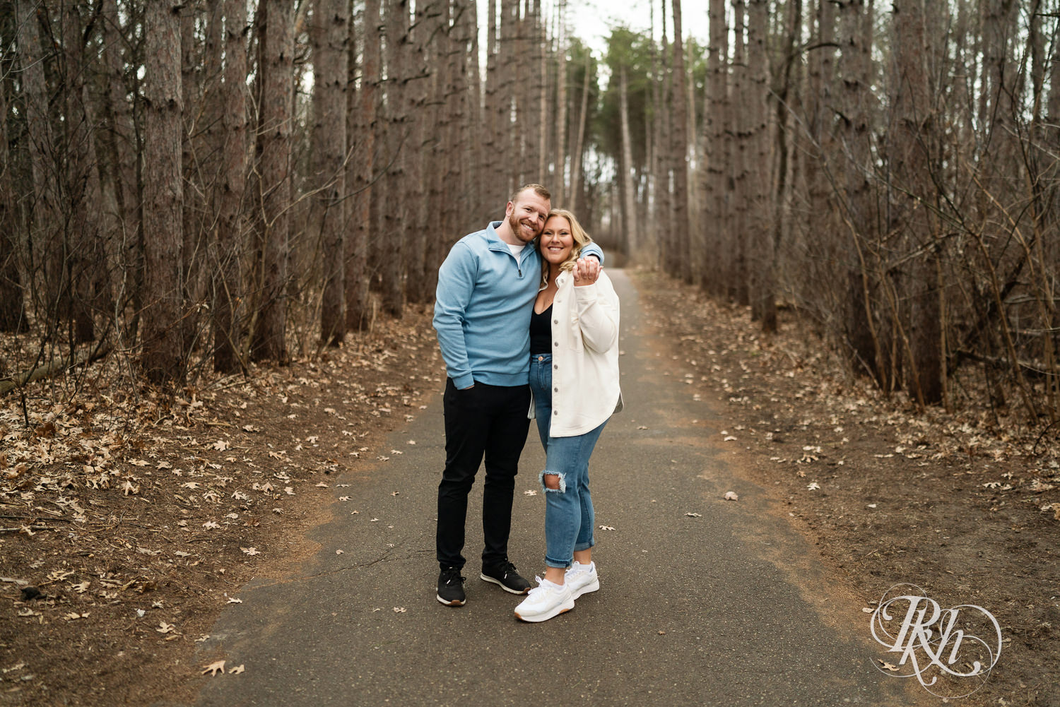 Man in blue shirt and woman in jeans smile between tall trees during engagement photography at Rice Creek Regional Trail in Shoreview, Minnesota.