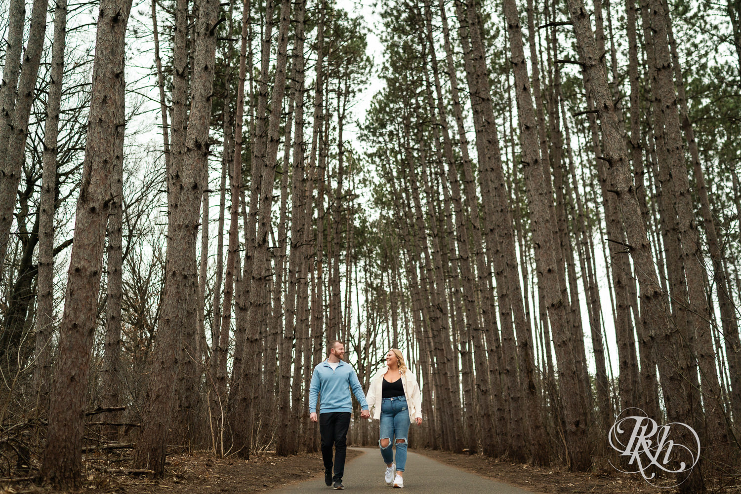 Man in blue shirt and woman in jeans walk between tall trees during engagement photography at Rice Creek Regional Trail in Shoreview, Minnesota.