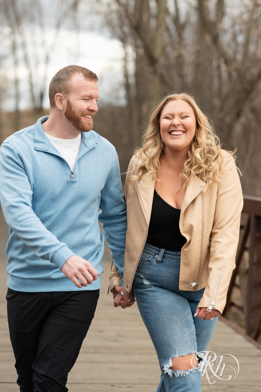 Man in blue shirt and woman in jeans laugh on bridge during engagement photography at Rice Creek Regional Trail in Shoreview, Minnesota.