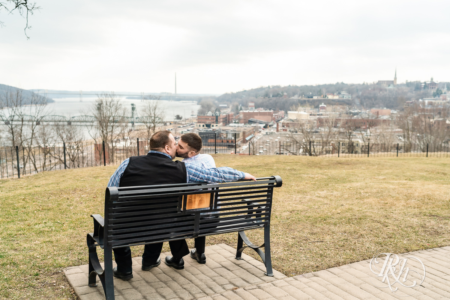 Two men in plaid shirts kiss on bench during engagement photography at Pioneer Park in Stillwater, Minnesota.