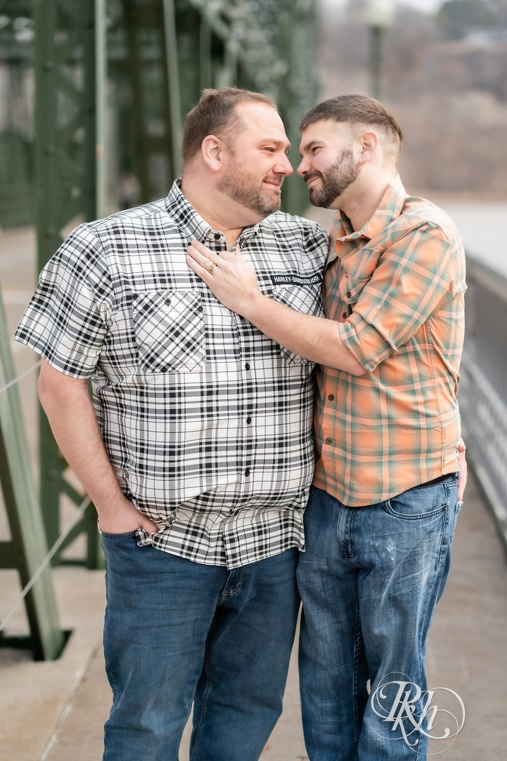 Two men in flannels and jeans kiss in on the bridge during engagement photography in Stillwater, Minnesota.