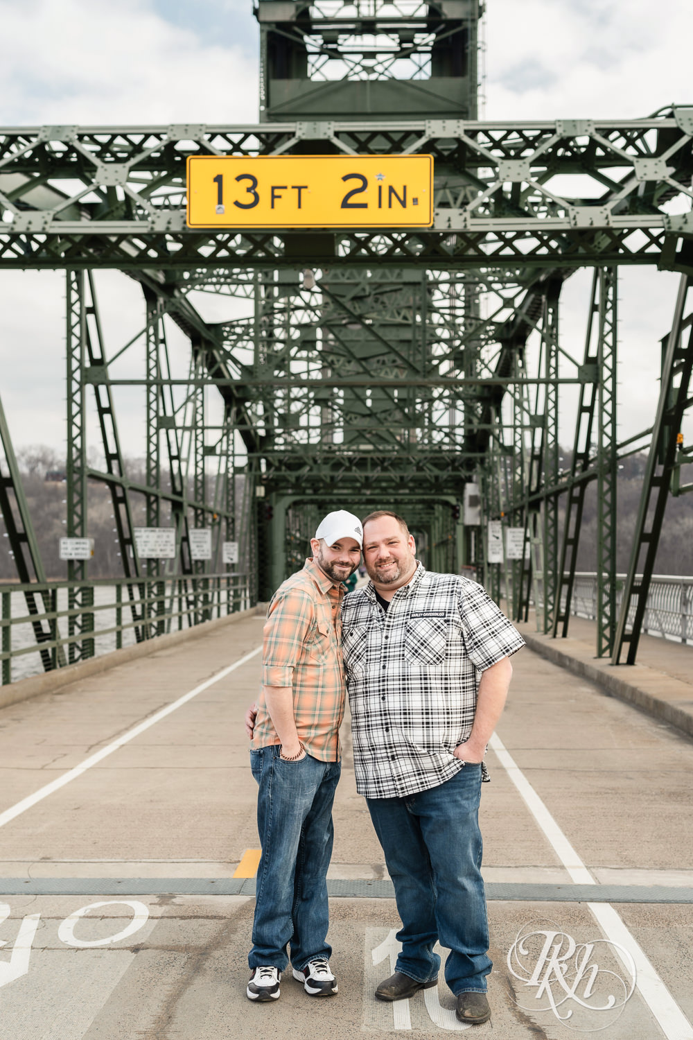 Two men in flannels and jeans smile on the bridge during engagement photography in Stillwater, Minnesota.