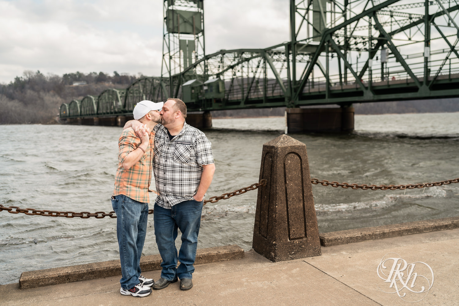 Two men in flannels and jeans kiss in front of the bridge during engagement photography in Stillwater, Minnesota.