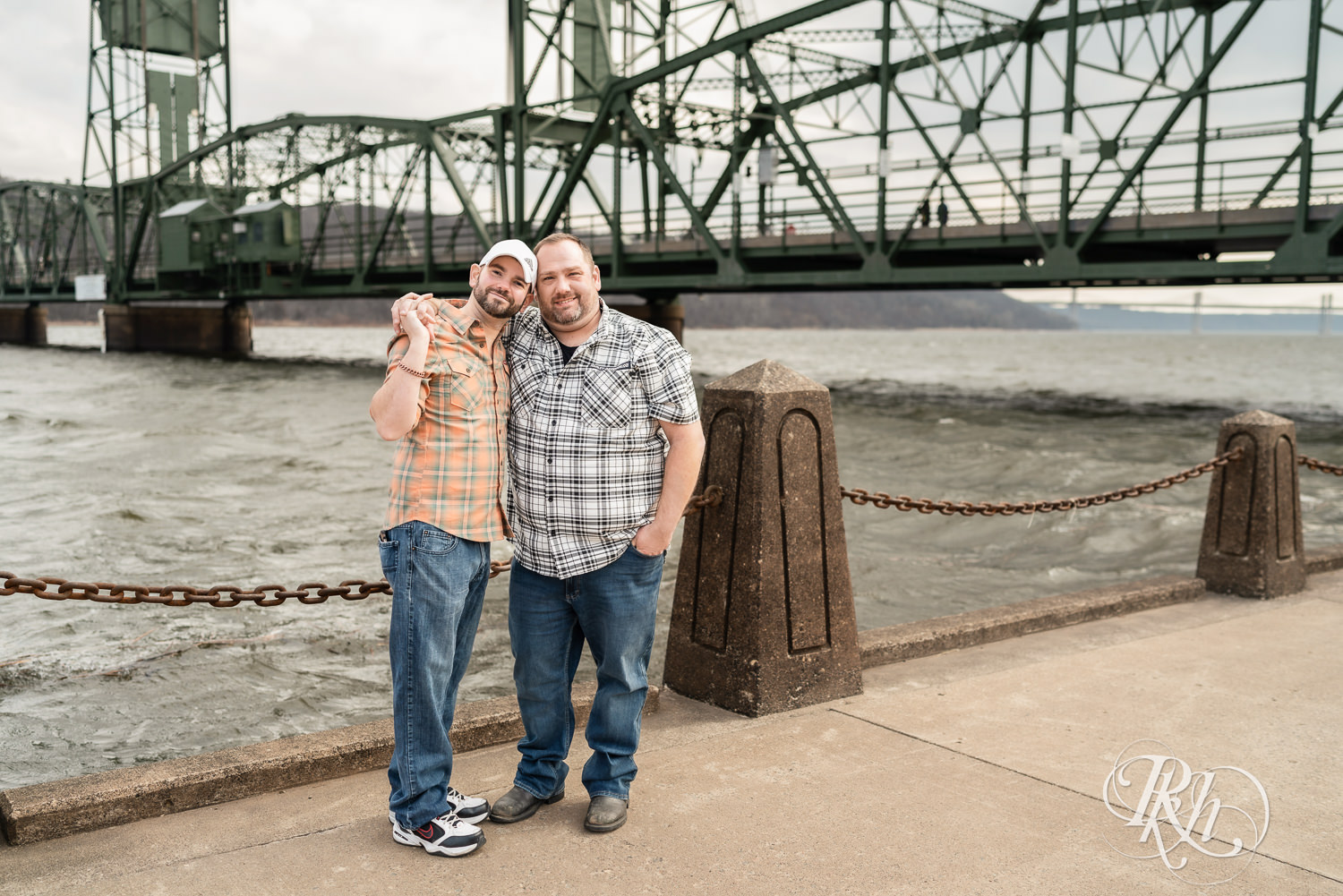 Two men in flannels and jeans smile in front of the bridge during engagement photography in Stillwater, Minnesota.