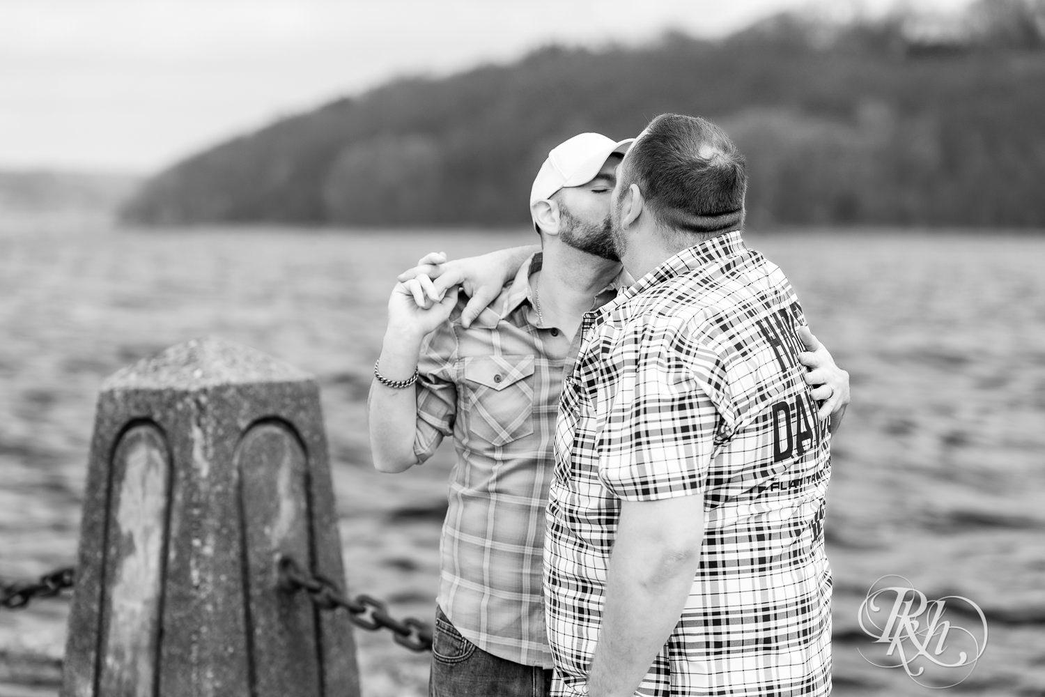 Two men in flannels and jeans kiss in front of the bridge during engagement photography in Stillwater, Minnesota.
