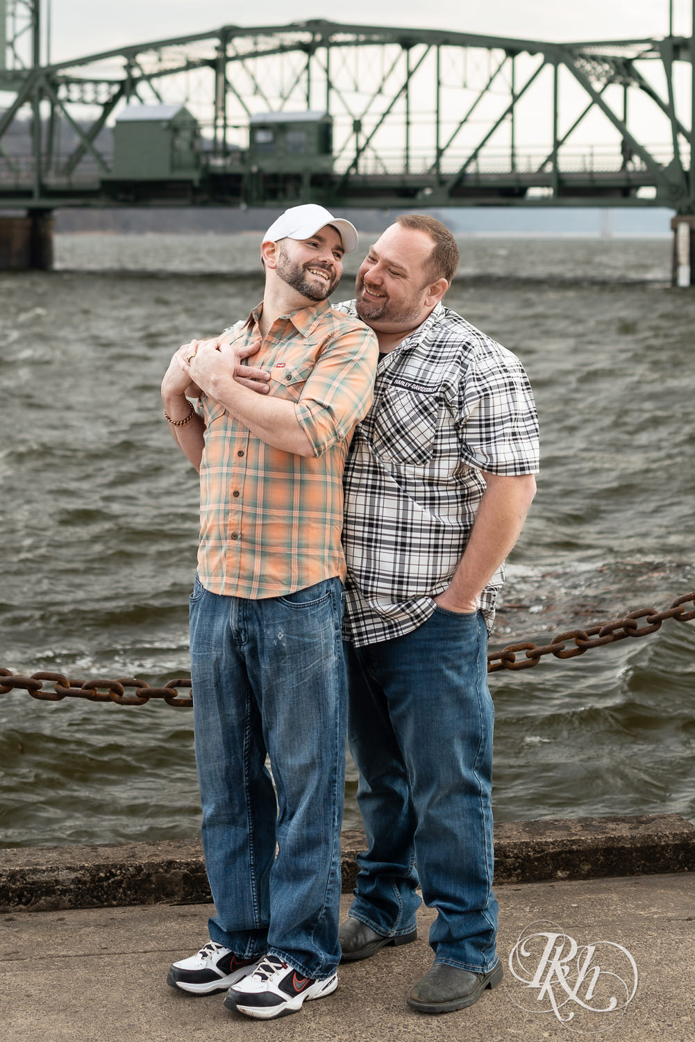 Two men in flannels and jeans smile in front of the bridge during engagement photography in Stillwater, Minnesota.