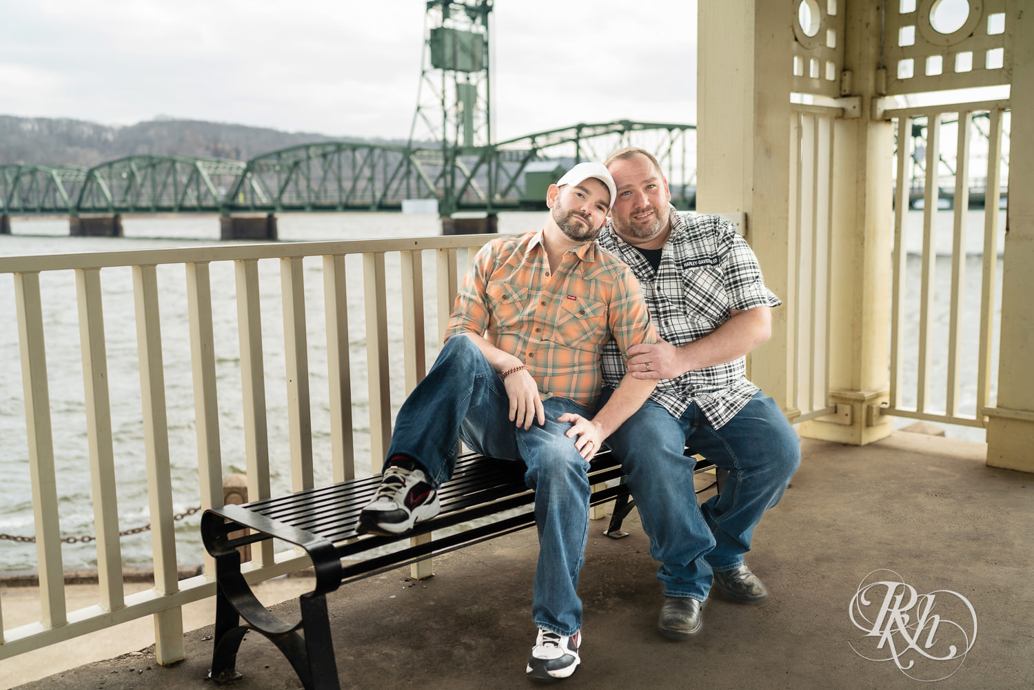 Two men in flannels and jeans smile on a bench during engagement photography in Stillwater, Minnesota.