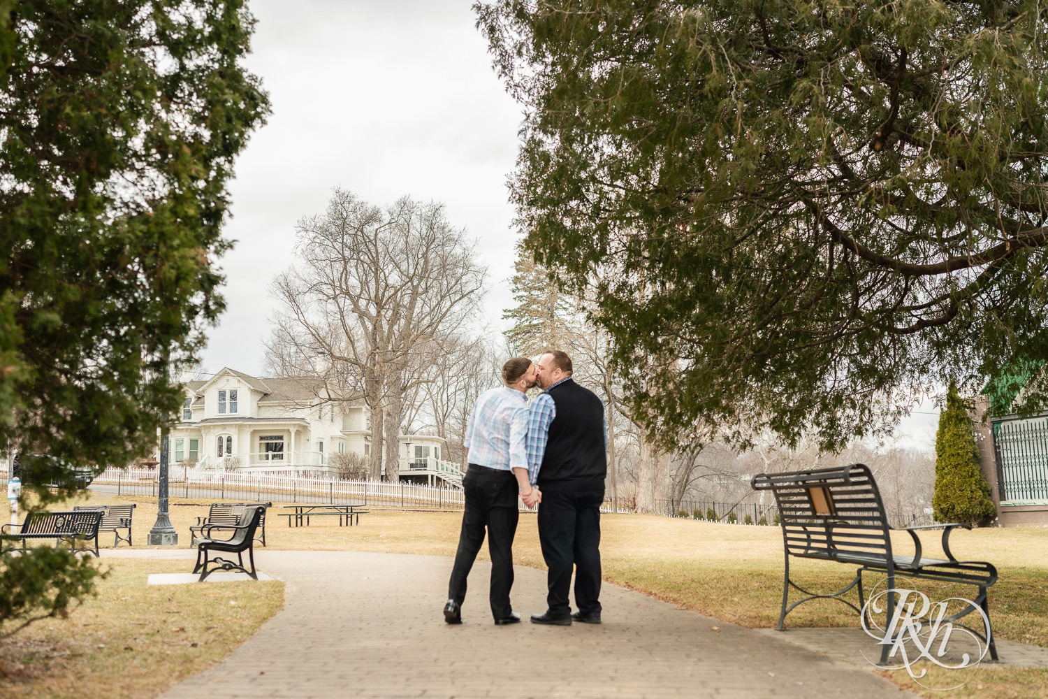Two men in plaid shirts kiss during engagement photography at Pioneer Park in Stillwater, Minnesota.