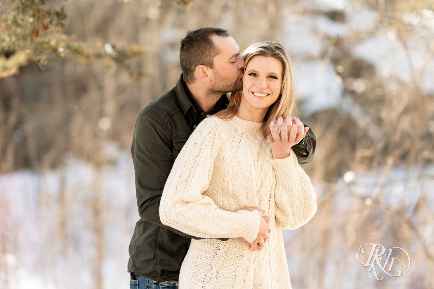 Man in jeans and green shirt and woman in a white sweater kiss in the snow at sunrise during Taylor's Falls engagement photography session.