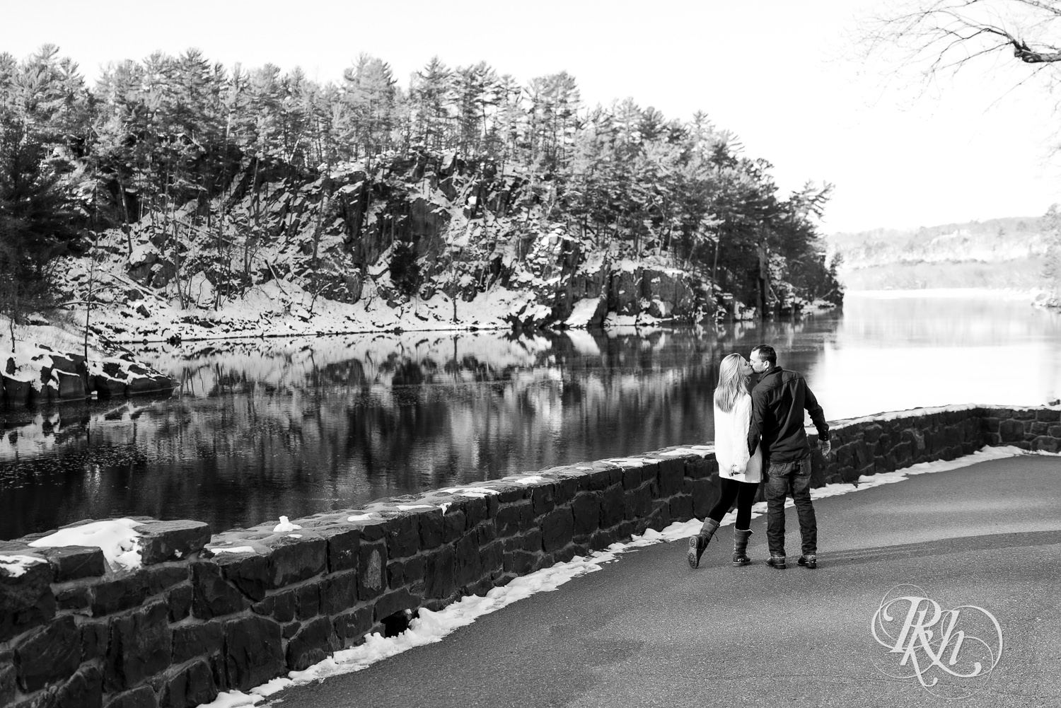 Man in jeans and green shirt and woman in a white sweater kiss while walking during Taylor's Falls engagement photography session.