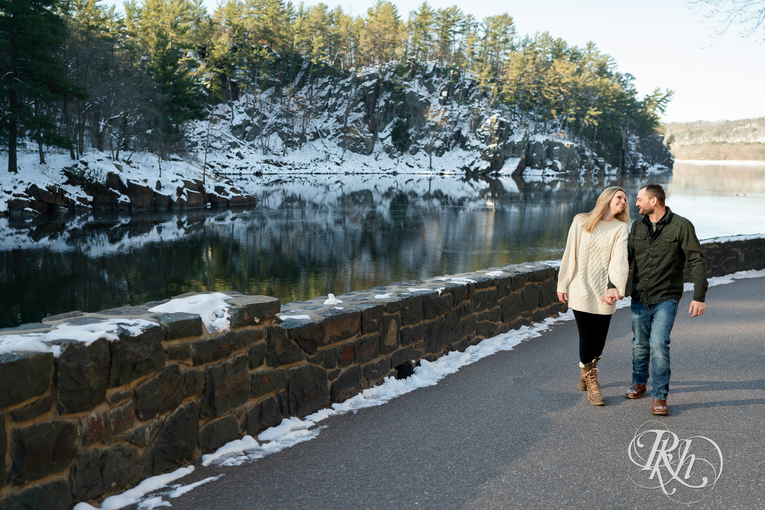Man in jeans and green shirt and woman in a white sweater smile while walking during Taylor's Falls engagement photography session.