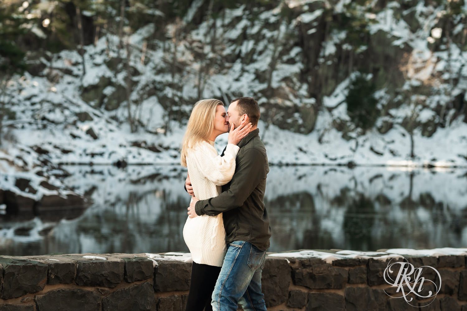 Man in jeans and green shirt and woman in a white sweater kiss in front of the river during Taylor's Falls engagement photography session.