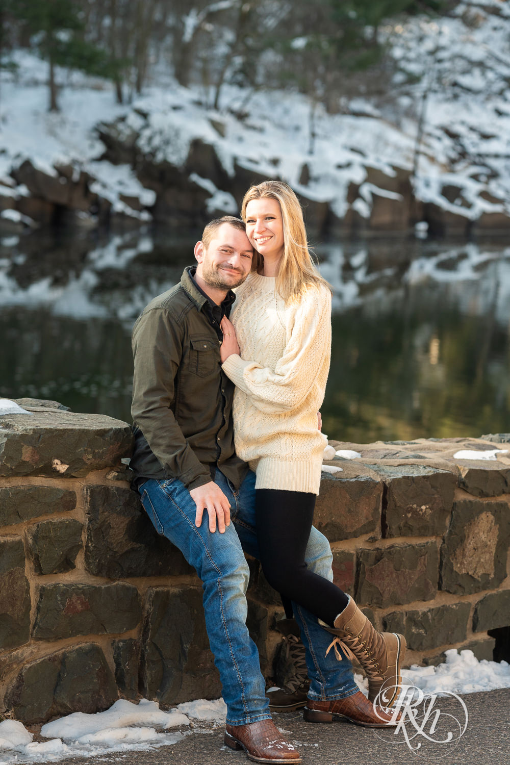 Man in jeans and green shirt and woman in a white sweater smile in front of the river at winter engagement photography session.