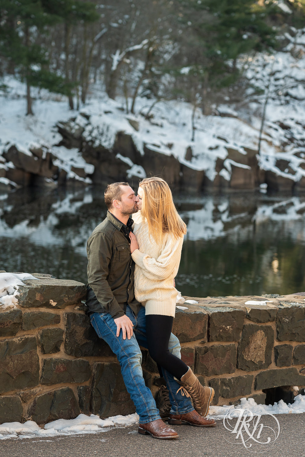 Man in jeans and green shirt and woman in a white sweater kiss in front of the river during Taylor's Falls engagement photography session.