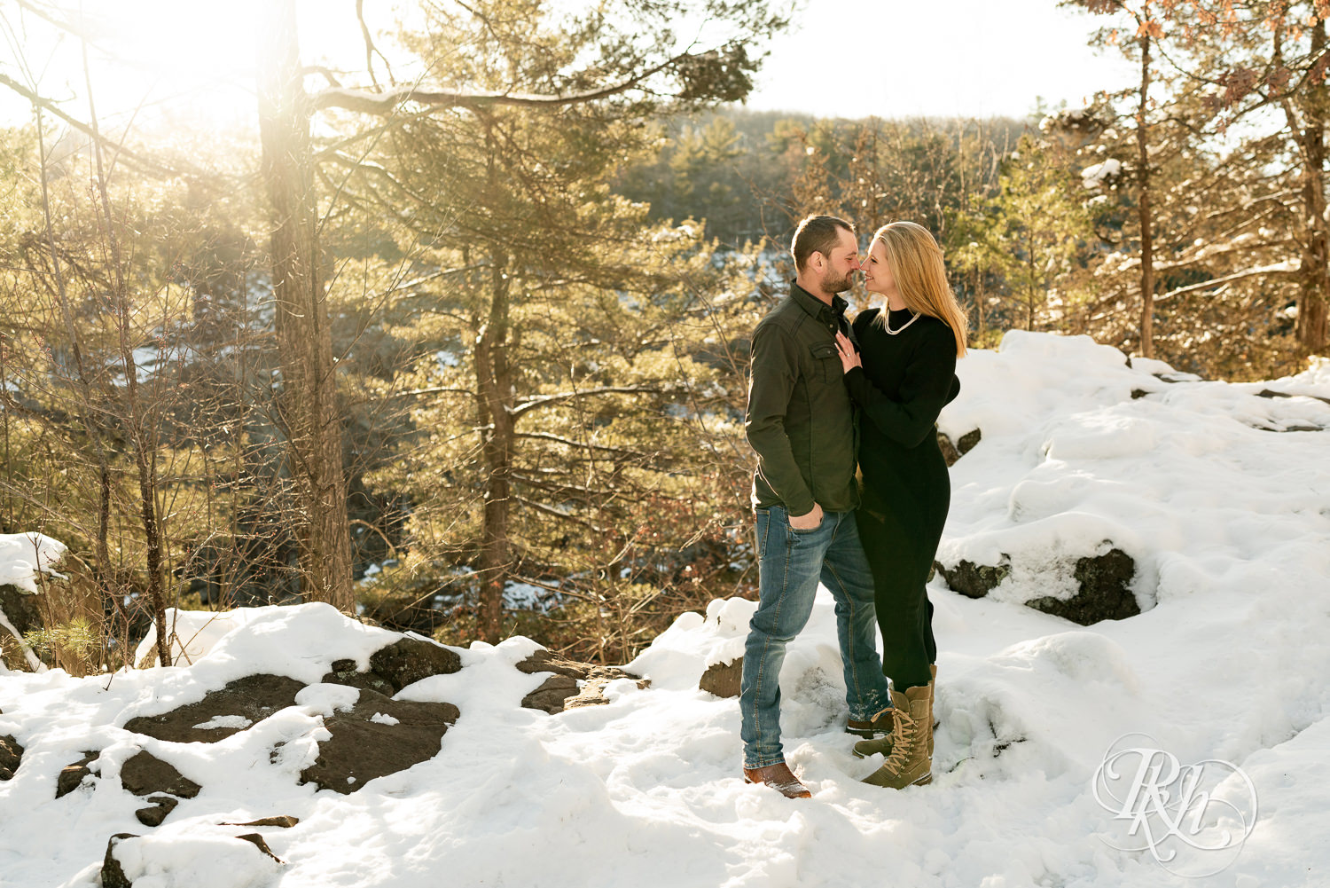 Man in jeans and woman in black dress smile in the snow at sunrise during Taylor's Falls engagement photography session.