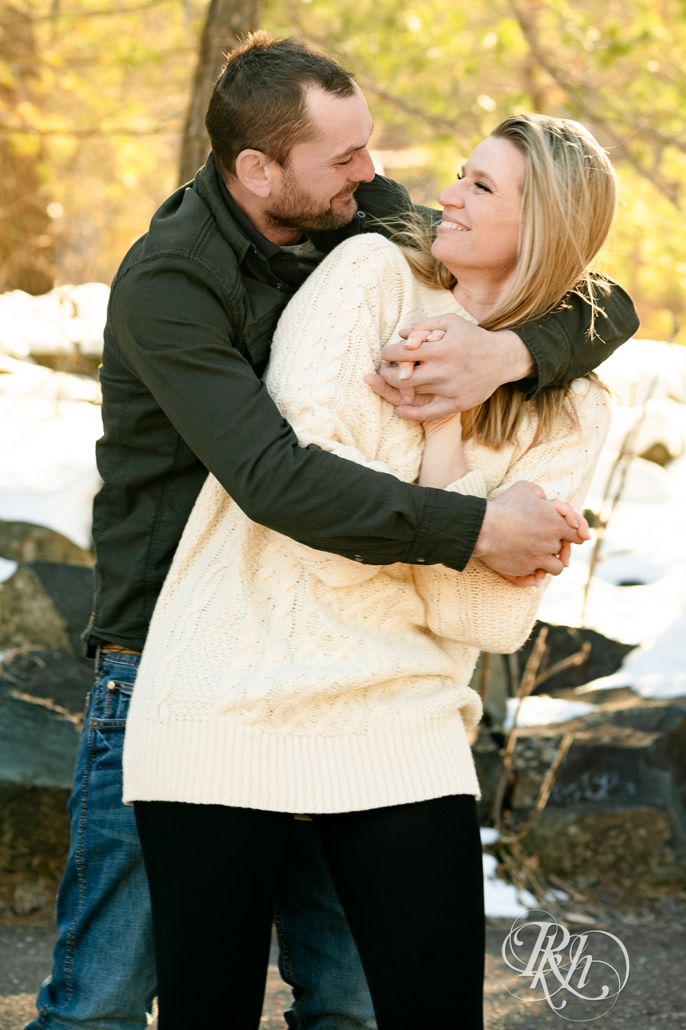 Man in jeans and green shirt and woman in a white sweater smile laugh at winter engagement photography session in Taylors Falls, Minnesota.